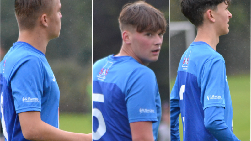 Three Basildon Town FC Under 17 players dressed in blue and showing RJS Waste Management sponsorship logos on their sleeves